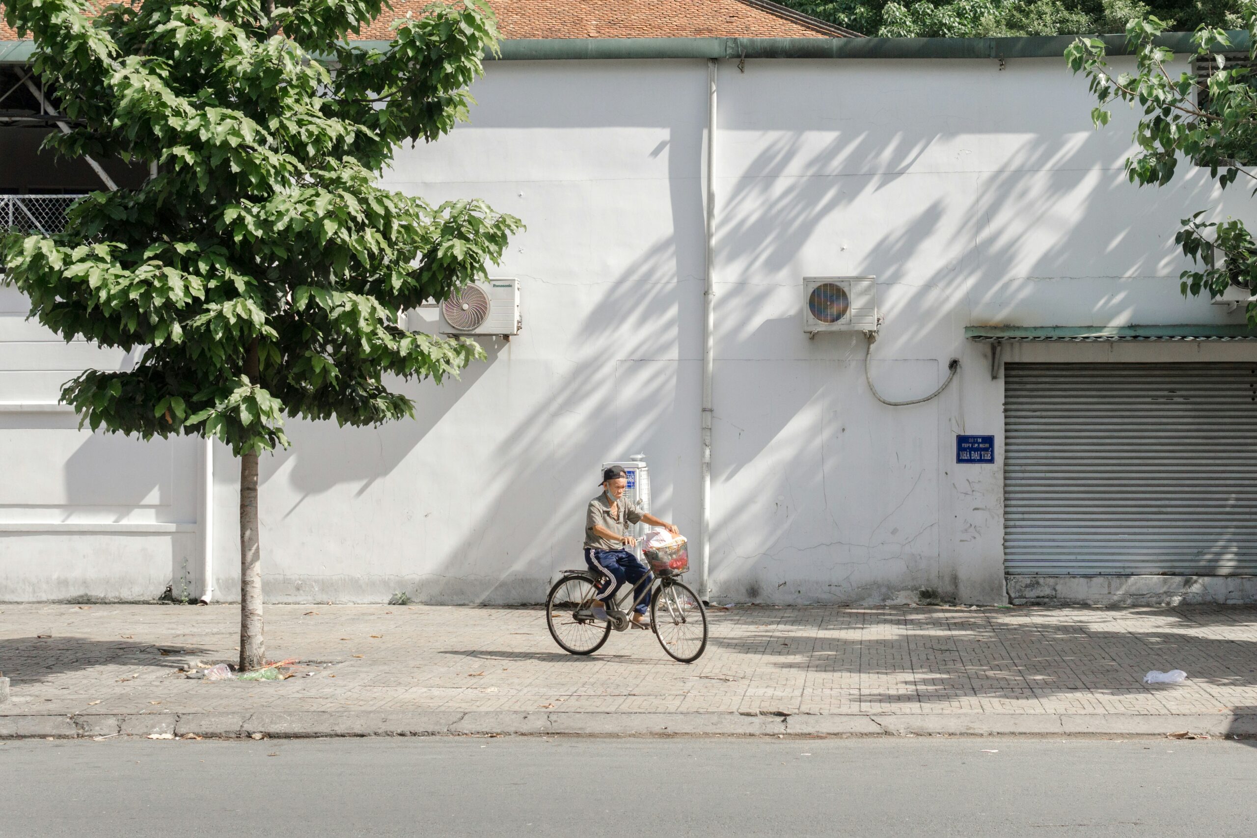 An elderly man riding a bicycle on a sidewalk in Saigon, captured by photographer Hiep Duong against a minimalist white building background.