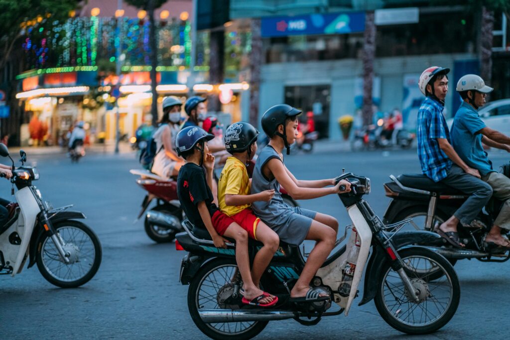 A candid, eye-level shot captures three young boys riding together on a single motorbike through a busy street intersection in Vietnam at dusk. The boy in the front steers the motorbike, while the other two sit behind him, all wearing helmets. In the background, blurry city lights, storefronts, and other motorbike riders create a lively urban atmosphere. Photo by @tronle_sg on Unsplash.